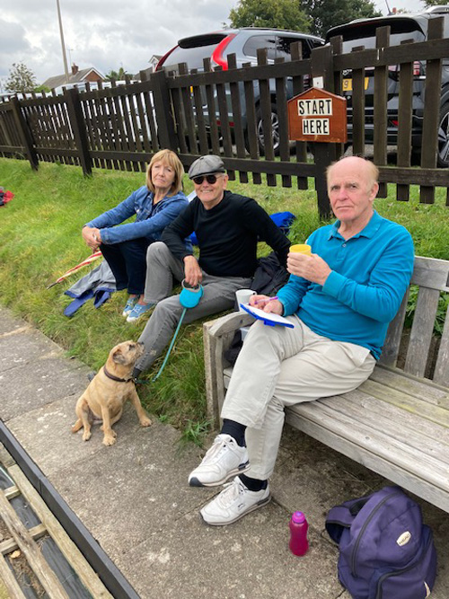 Laura, Bill (with Sally the dog) sitting on the grass and Mike sitting on a bench, scoring. At the Merit.