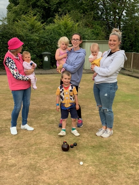 Three women with babies standing on the green with small child in front of them