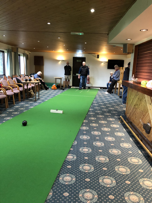 Bowlers standing and sitting to the side of the mat watching a wood which has just been delivered. Where will it end up?