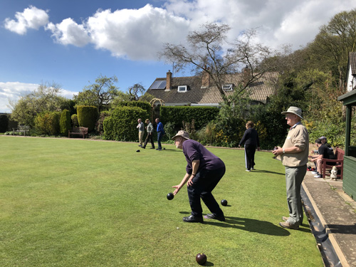 In the foreground a man bowls watched by his opponent. Ini the background, other games going on