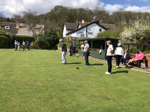 Groups of bowlers on the green, watched by spectators sitting on a bench