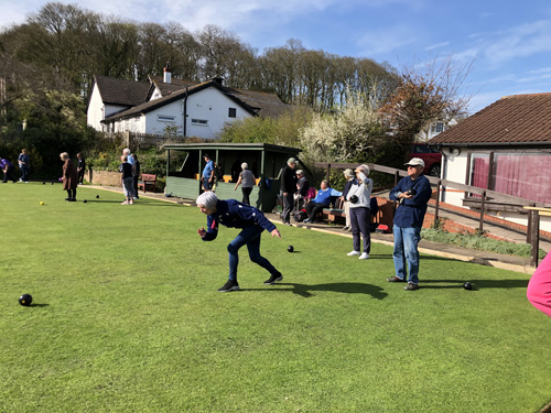 Margaret bowls watched by Alan. Other games going on in the background. Spectators sitting on benches around the green