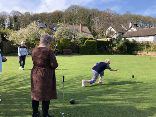 Brenda bowls watched by Wendy. People walking around the green in the background