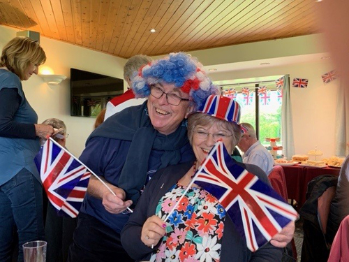 Alan and Wendy hugging and waving flags. Alan is wearing a red, white and blue curly wig. Wendy is wearing a Union Jack bowler.