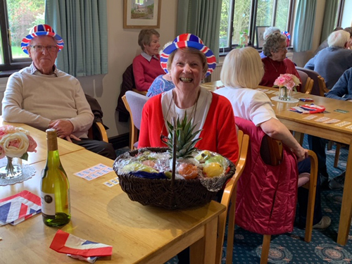 Joan and Brian at the table, both wearing Union Jack bowlers. A basket of fruit is in front of Joan.