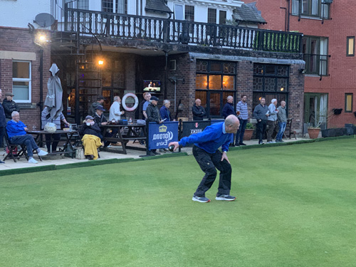 Geoff bowling at St Werburgh's, watched by home and away players, standing and sitting, drinking.