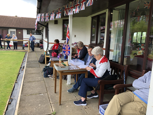 Pat & Ken working out who will be playing next. Sitting on the bench under the Clubhouse eaves. Other bowlers sitting watching the games. Union Jack bunting all along the outside of the Clubhouse.