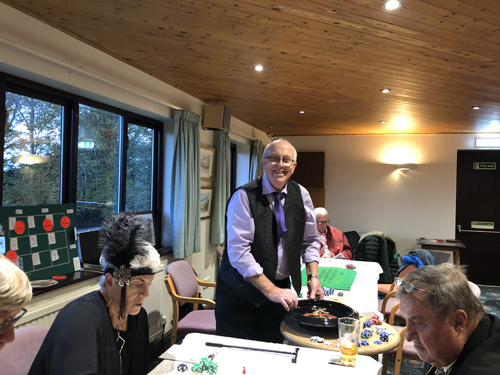 Alan stands by the roulette wheel, smiling. People in foreground intent on where their bets are.