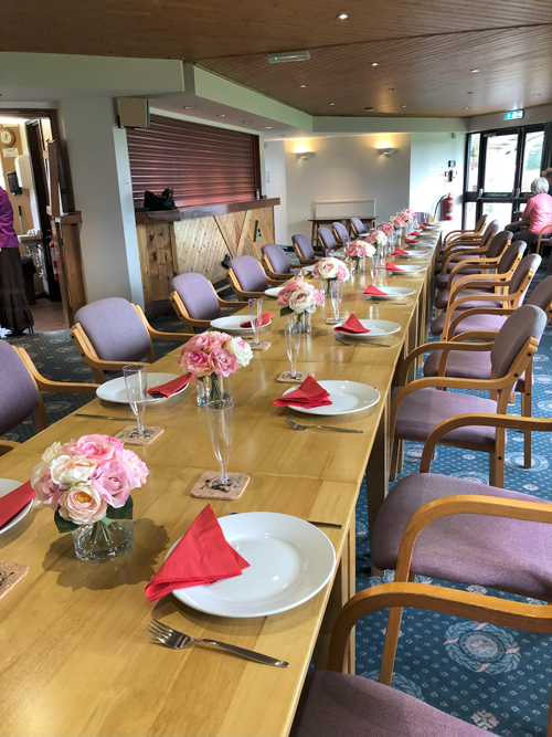 Picture of the Clubhouse with long table set with vases of flowers down the centre and place settings.