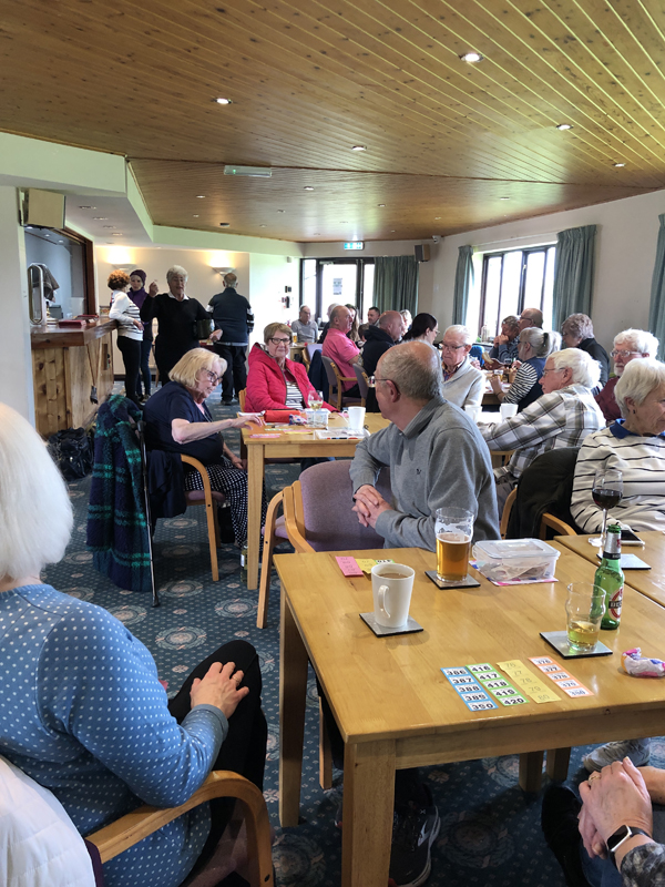 The raffle drawing in progress. Taken from the back of the Clubhouse towards the french doors. Tables with people turned towards Pat who is drawing the raffle.