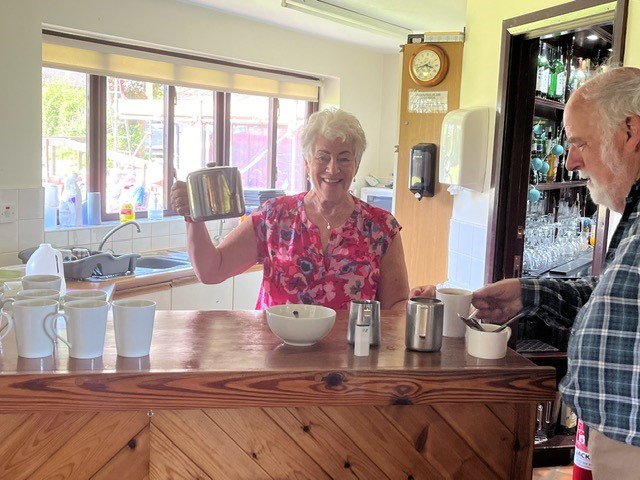 Pat H behind the counter serving teas. Holding up the teapot and smiling