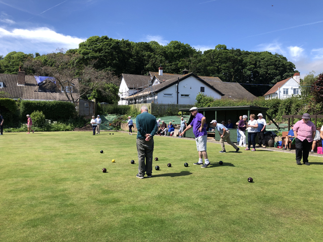 Bowlers on and around the green. Foreground two men are working out which bowls are nearest the jack.