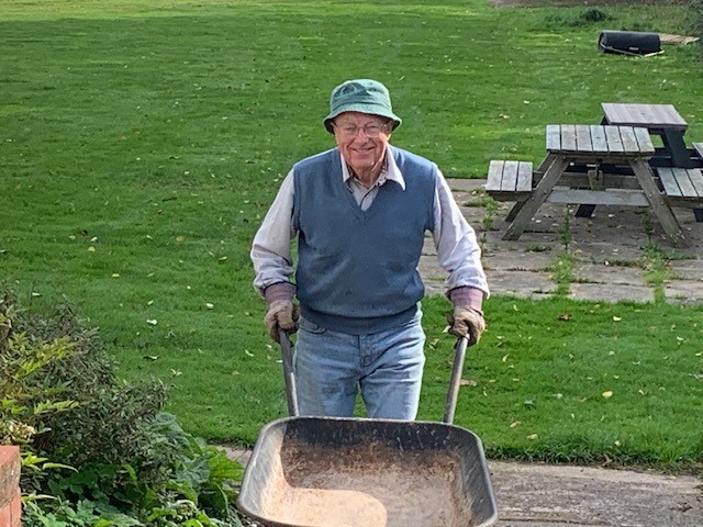 Geoff returns from the compost-heap with an empty wheelbarrow. He is smiling broadly.