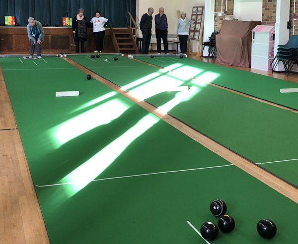 Three Indoor Bowls mats in a church hall. Players at the far end. On the left hand side, a woman is bowling towards four bowls.