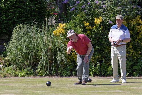 Two men on the green. One has just bowled and the other is standing behind him watching