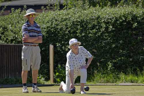 A man and a woman on the green. The woman is one knee, having just bowled. The man is standing behind her watching.